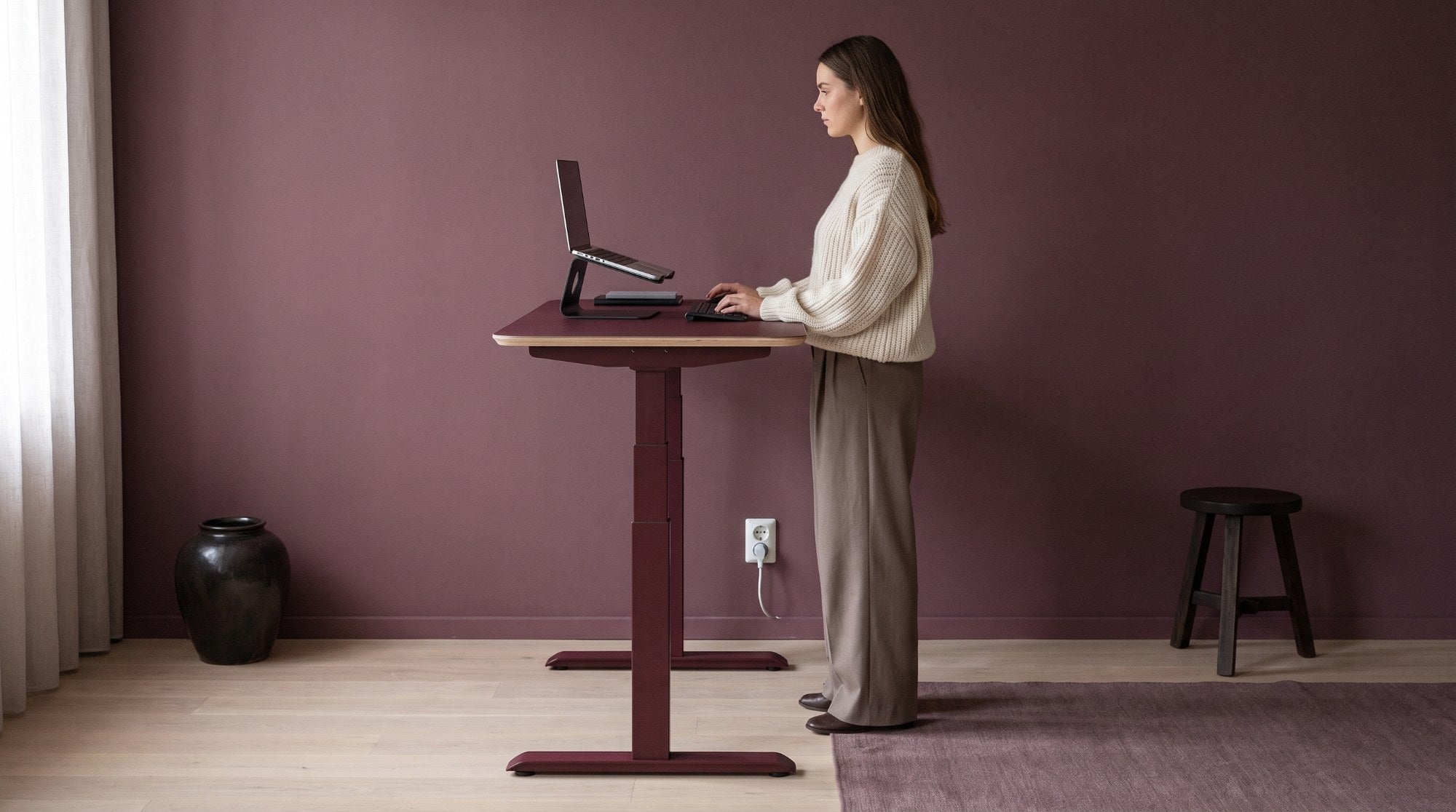 Vrouw werkt staand aan een Stadesk One in burgundy met frame in dezelfde kleur, tegen een pruimkleurige wand